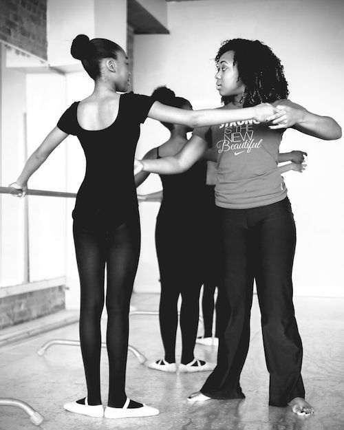 Karisma Jay helps a student at the ballet barre. The students wear black leotards and tights and stand in first position. Jay helps a student lengthen her arms.